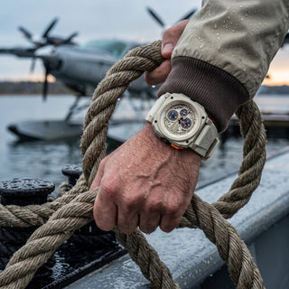 A close-up lifestyle shot of a person's hand holding a thick rope on a boat deck, wearing a beige revolution watch with a white dial and orange accents. A waterplane airplane is visible in the blurred background over the water.