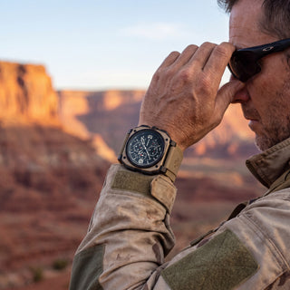 A man in outdoor gear wearing a rugged tactical revolution watch on his wrist, with a desert canyon landscape blurred in the background.