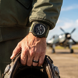 A close-up of a man's hand wearing an olive green military-style watch with a black dial, holding a vintage leather helmet with a blurred propeller plane in the background.