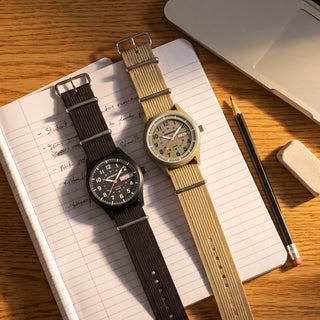 Two analog field watches with nato straps are arranged on an open lined notebook on a wooden desk, with a laptop corner, pencil, and eraser nearby. One watch has a black dial and dark brown strap, and the other has a beige dial and tan strap. back to school field watch REVO-FW01